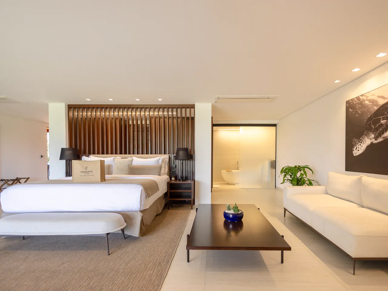 Suíte de resort com cama de casal arrumada, sofá claro e banheiro ao fundo.